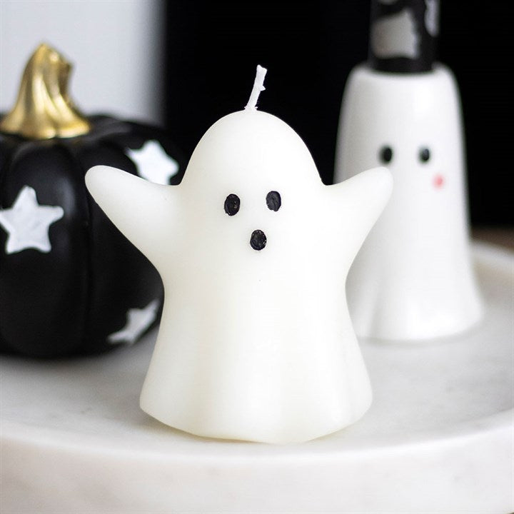 Ghost-shaped candle with black eyes and mouth on a white surface with Halloween decorations.